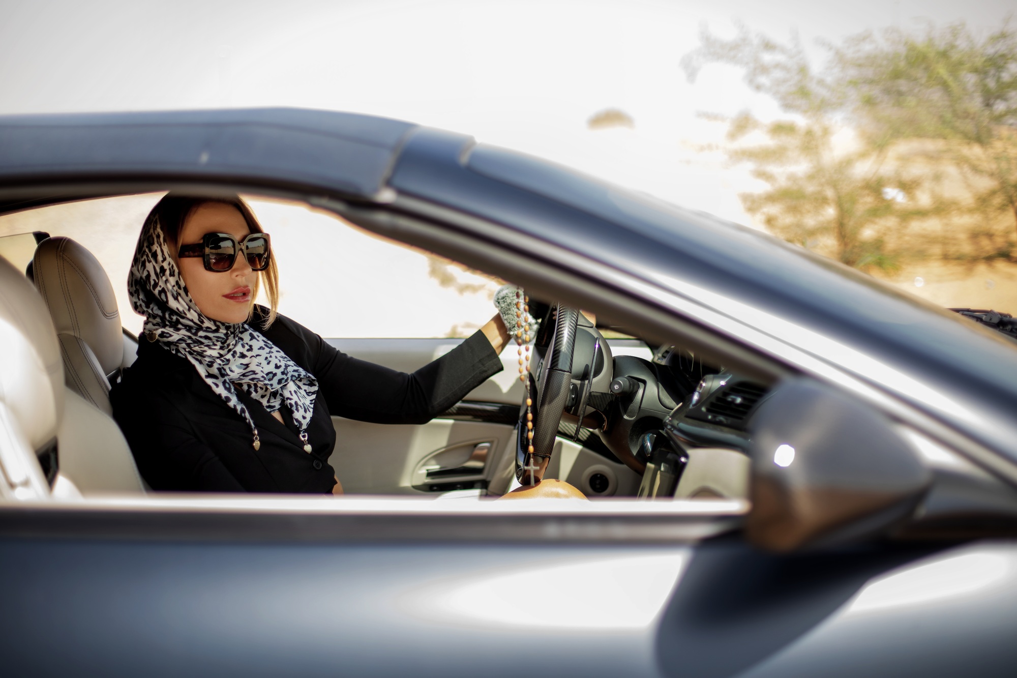 A fashionable modern woman in sunglasses and a silk scarf sits in a premium car in the desert, Dubai